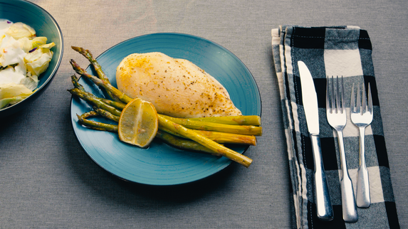 Plate of Mediterranean Garlic Roasted Chicken served with roasted asparagus and lemon slices on a blue plate, accompanied by a side salad in a bowl, with cutlery set on a checkered napkin on a grey tablecloth.